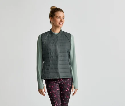Une femme portant un chignon porte une veste sans manches matelassée grise sur un t-shirt à manches longues vert clair et un legging à motifs.