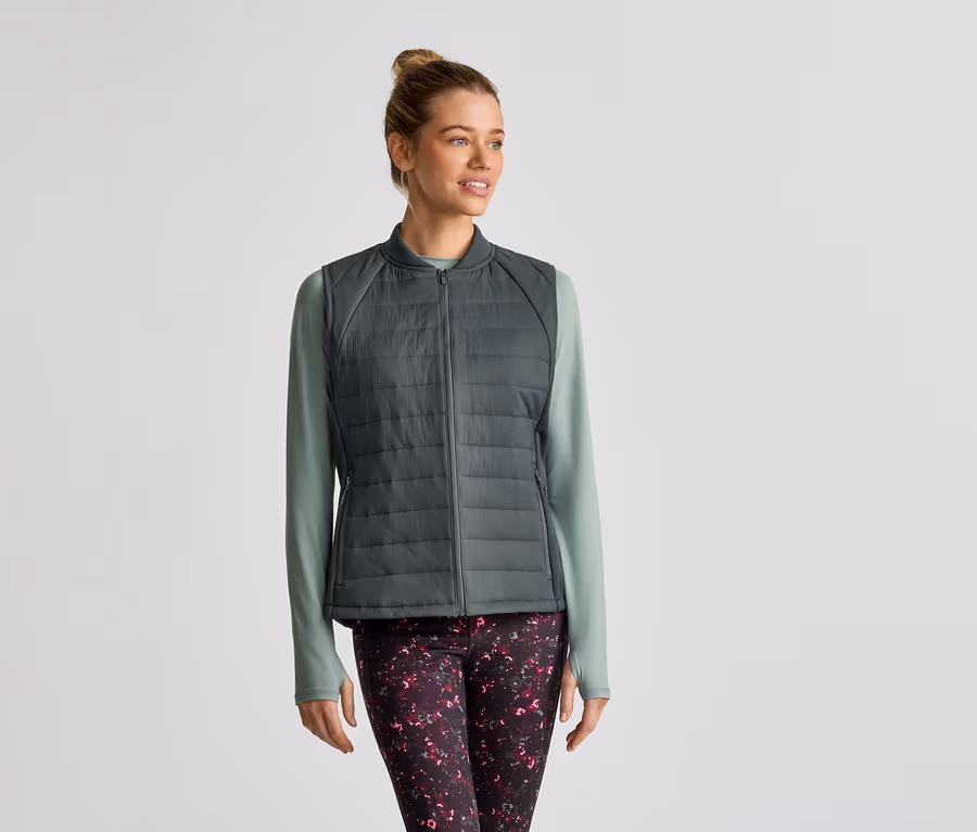 Une femme portant un chignon porte une veste sans manches matelassée grise sur un t-shirt à manches longues vert clair et un legging à motifs.