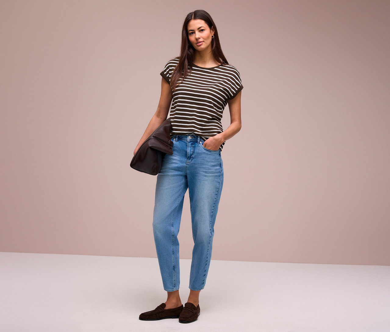 Une femme aux longs cheveux foncés est debout, regardant devant elle. Elle porte un T-shirt Street One rayé, un jean bleu clair et des mocassins marron foncé, tenant un sac marron sous son bras gauche.