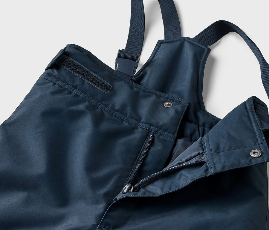 Gros plan d'un pantalon de pluie bleu marine avec bretelles, fermeture éclair partiellement ouverte et bouton-pression argenté.