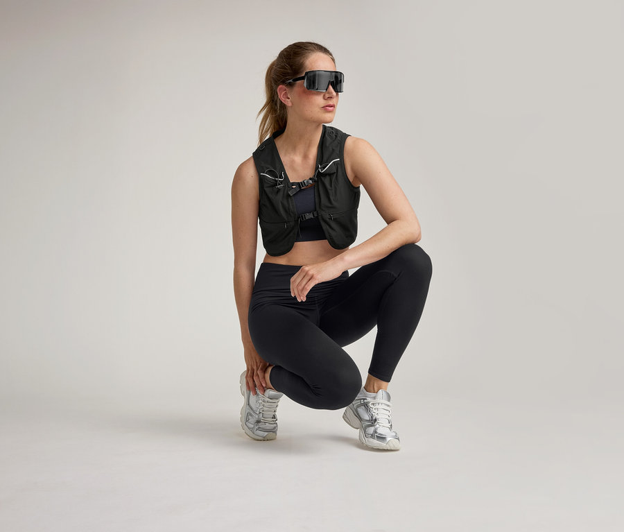 Femme en tenue de sport noire, gilet et lunettes de soleil accroupie sur fond clair.