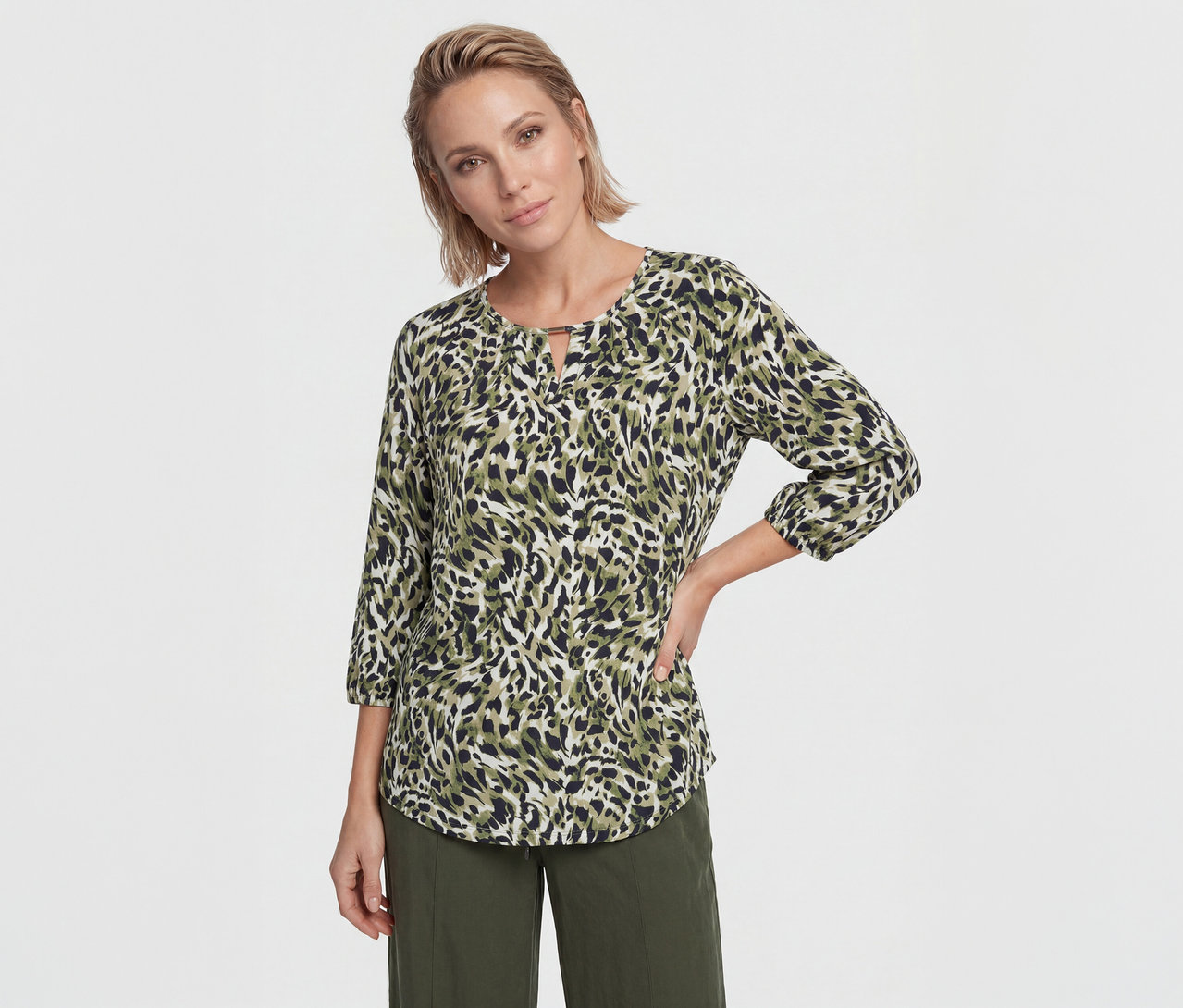 Femme aux cheveux blonds courts portant un chemisier à imprimé animal vert, noir et blanc avec des manches trois-quarts et un pantalon vert olive, posant la main sur la hanche.