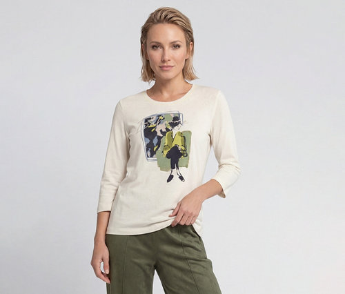 Femme blonde portant un t-shirt crème à manches longues avec un imprimé graphique d'une femme en veste jaune et un pantalon vert olive.