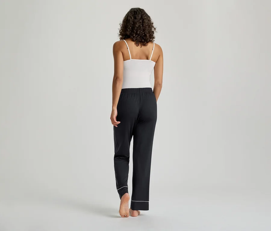Une femme est debout, de dos, vêtue d'un débardeur blanc à fines bretelles et d'un pantalon de pyjama noir avec un passepoil blanc aux chevilles. Ses cheveux foncés et bouclés sont visibles, et elle est pieds nus sur un fond gris clair.