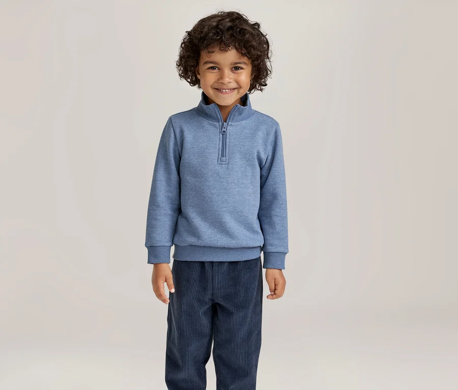 Garçon souriant aux cheveux bouclés, portant un sweat-shirt bleu à demi-zip et un pantalon bleu foncé.