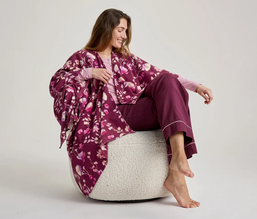 Une femme souriante aux longs cheveux bruns est assise pieds nus sur un pouf blanc moelleux, vêtue d'un haut rose pâle à manches longues, d'un pantalon large bordeaux avec une bordure rose et d'une robe de chambre kimono bordeaux à motifs floraux.