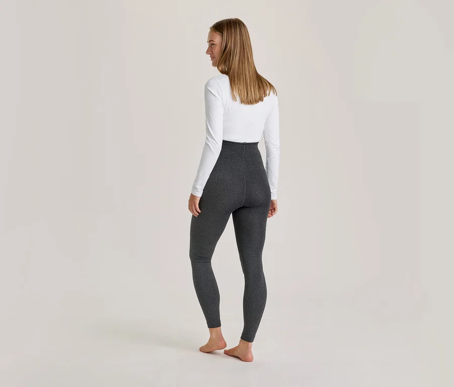 Femme debout de dos, portant un haut blanc à manches longues et un legging gris foncé, pieds nus sur un fond clair.