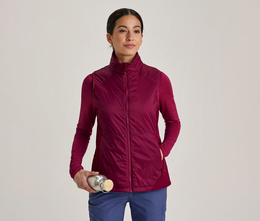 Femme portant un gilet matelassé bordeaux sur un t-shirt à manches longues et un pantalon bleu, tenant une bouteille d'eau argentée avec un bouchon en bois.