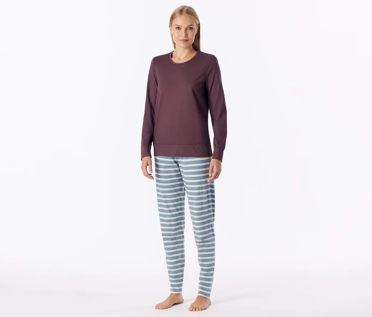 Femme debout portant un haut à manches longues violet foncé et un pantalon de pyjama rayé bleu clair et blanc, pieds nus.