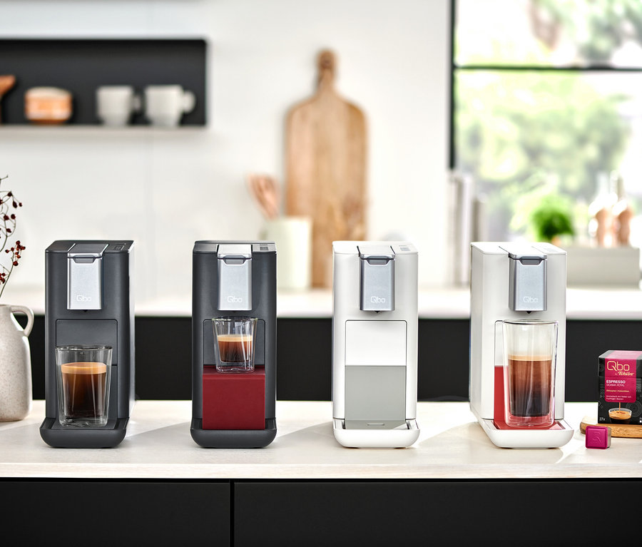 Quatre machines à café Qbo ESSENTIAL en Dark Berry, Dark Stone, Bright Coral et Bright Sand sont posées avec des expressos sur un comptoir de cuisine.