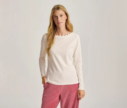 Femme aux longs cheveux blonds portant un haut à manches longues crème avec un col festonné et un pantalon en velours côtelé rose, une main dans la poche.
