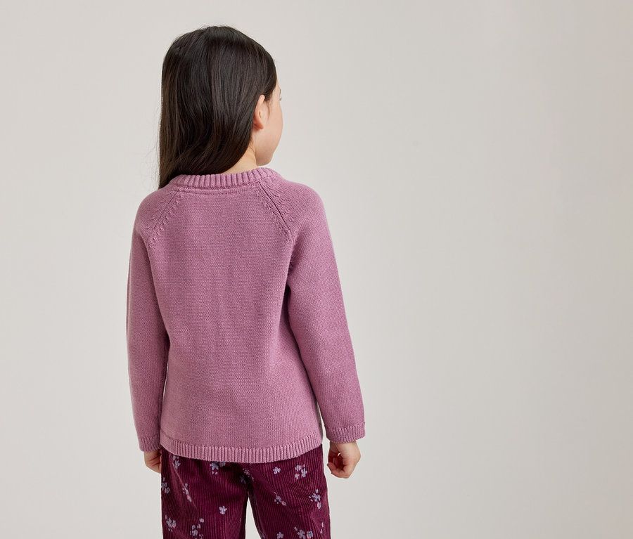 Une jeune fille vue de dos, portant un pull rose à manches longues et un pantalon rouge foncé à motif floral clair.