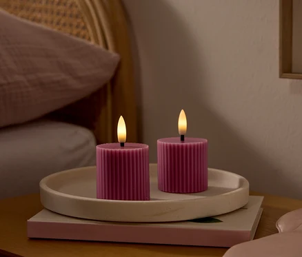 Deux bougies LED roses et côtelées avec des mèches lumineuses sont posées sur un plateau clair sur une table de chevet en bois. Un masque de sommeil rose est à côté. À l'arrière-plan, un lit avec une tête de lit en rotin et un oreiller rose.