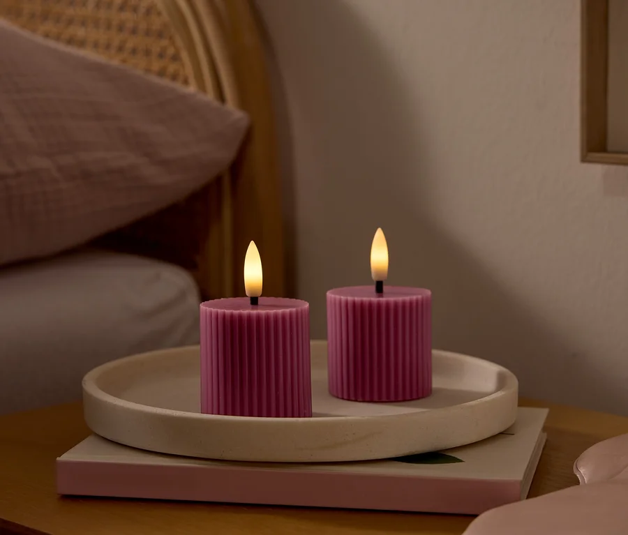 Deux bougies LED roses et côtelées avec des mèches lumineuses sont posées sur un plateau clair sur une table de chevet en bois. Un masque de sommeil rose est à côté. À l'arrière-plan, un lit avec une tête de lit en rotin et un oreiller rose.