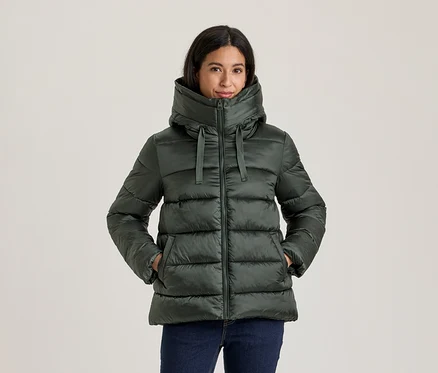Femme portant une doudoune vert foncé avec un col montant et une capuche, mains dans les poches, avec un jean bleu foncé.