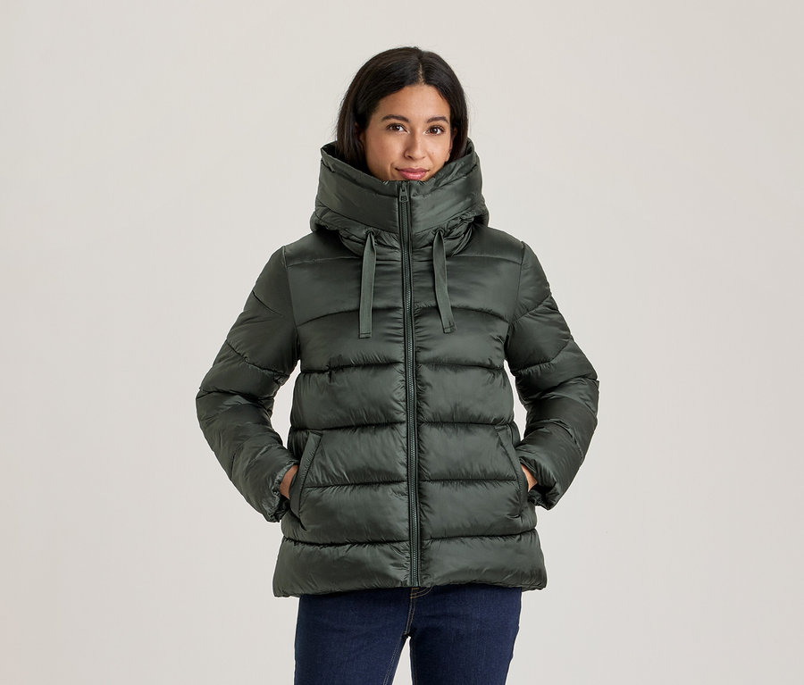 Femme portant une doudoune vert foncé avec un col montant et une capuche, mains dans les poches, avec un jean bleu foncé.