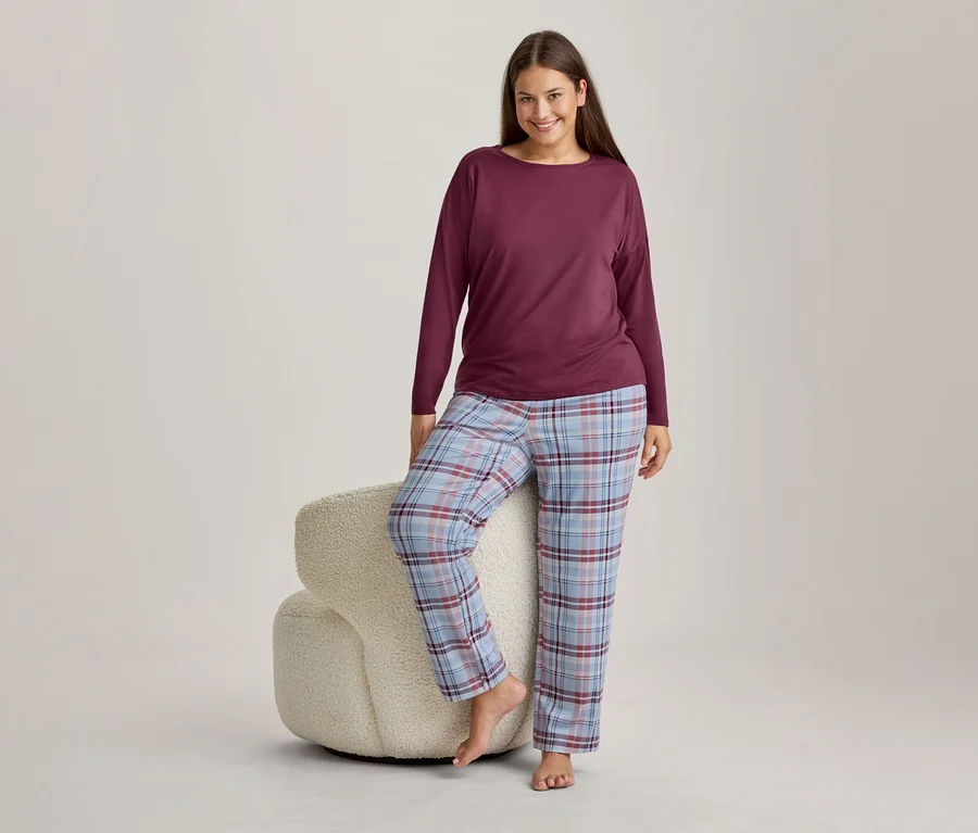Femme souriante portant un haut bordeaux à manches longues et un pantalon de pyjama à carreaux bleu clair, rouge et blanc, debout avec un pied sur un tabouret blanc moelleux.
