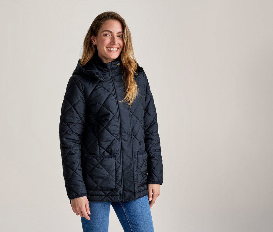 Femme souriante aux cheveux longs portant une veste matelassée bleu foncé à capuche et un jean bleu.