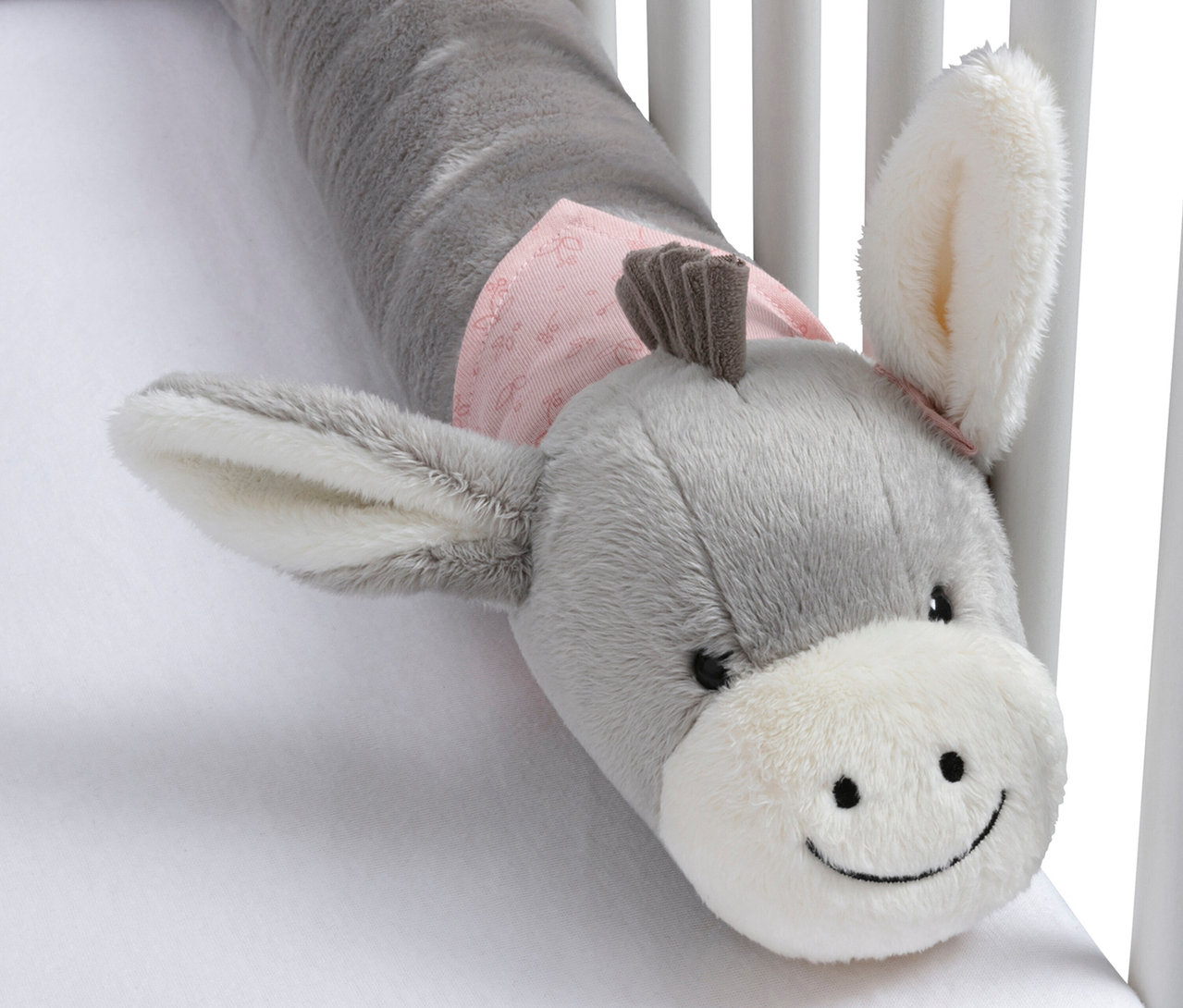 Pare-chocs de lit en peluche grise en forme d'âne avec un museau blanc et un tissu rose autour du cou, posé sur un drap blanc dans un lit bébé.