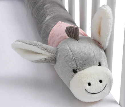 Pare-chocs de lit en peluche grise en forme d'âne avec un museau blanc et un tissu rose autour du cou, posé sur un drap blanc dans un lit bébé.
