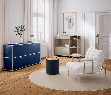 Un salon moderne avec un parquet en bois et des murs blancs. À gauche, un buffet modulaire bleu marine sous une fenêtre, avec un vase et une lampe. Au centre, un tapis rond avec une table d'appoint cylindrique et une bouteille d'eau. À droite, un fauteuil clair et une étagère roulante beige avec de la vaisselle. Un tableau abstrait est accroché au mur.