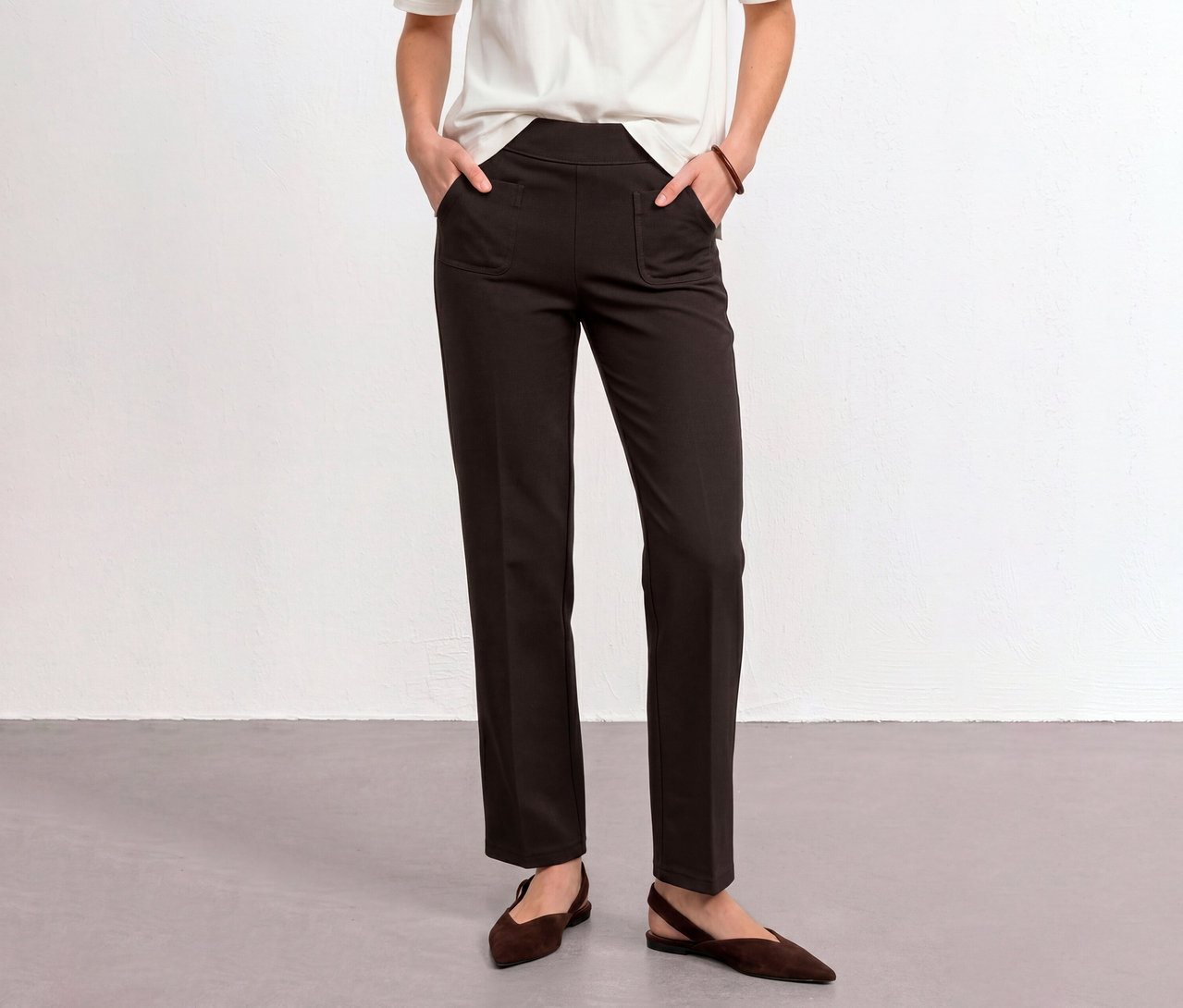 Une personne porte un pantalon en tissu Street One marron, les mains dans les poches, un t-shirt blanc et des chaussures plates marron.