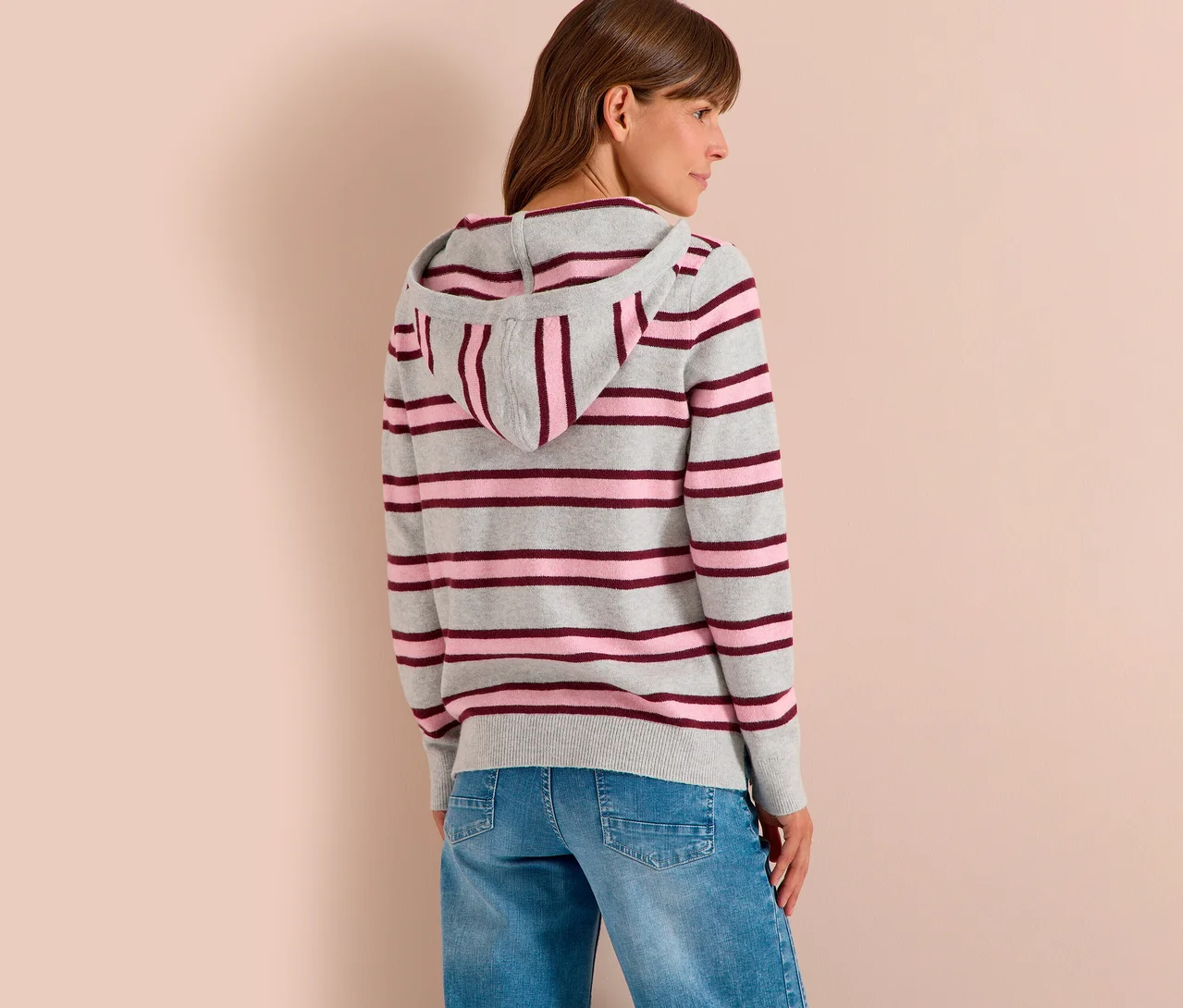 Femme de dos, légèrement tournée sur le côté, portant un sweatshirt à capuche en maille CECIL rayé gris, rose et bordeaux, et un jean bleu.