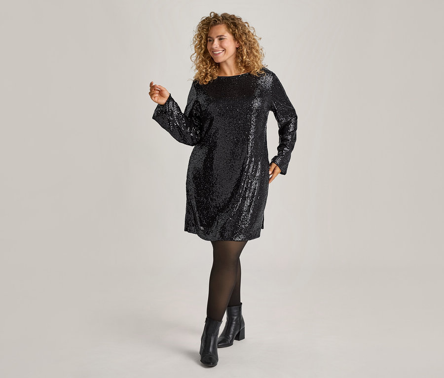 Femme souriante aux cheveux bouclés portant une mini-robe noire à paillettes, des collants fins doublés noirs en aspect transparent et des bottines noires.
