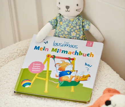 Livre « Leo Lausemaus – mein Mitmachbuch » avec une peluche de chat et d'autres jouets sur une surface duveteuse.