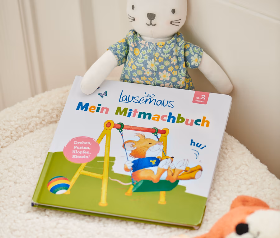 Livre « Leo Lausemaus – mein Mitmachbuch » avec une peluche de chat et d'autres jouets sur une surface duveteuse.