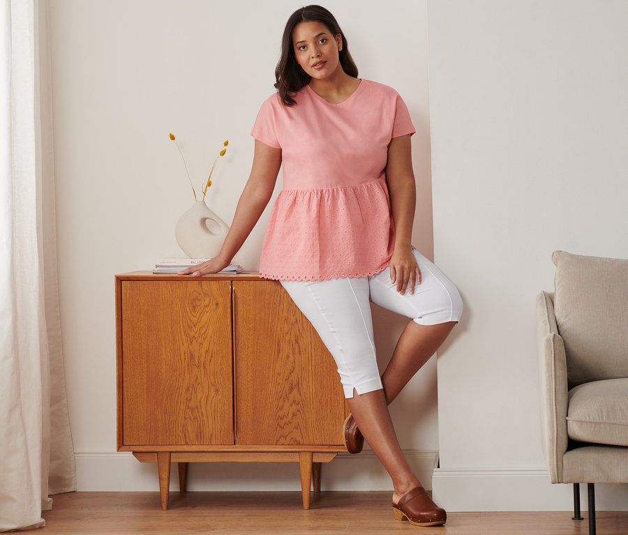 Femme appuyée sur une commode portant un t-shirt bimatière à broderie corail, un corsaire stretch blanc et des sabots en cuir.