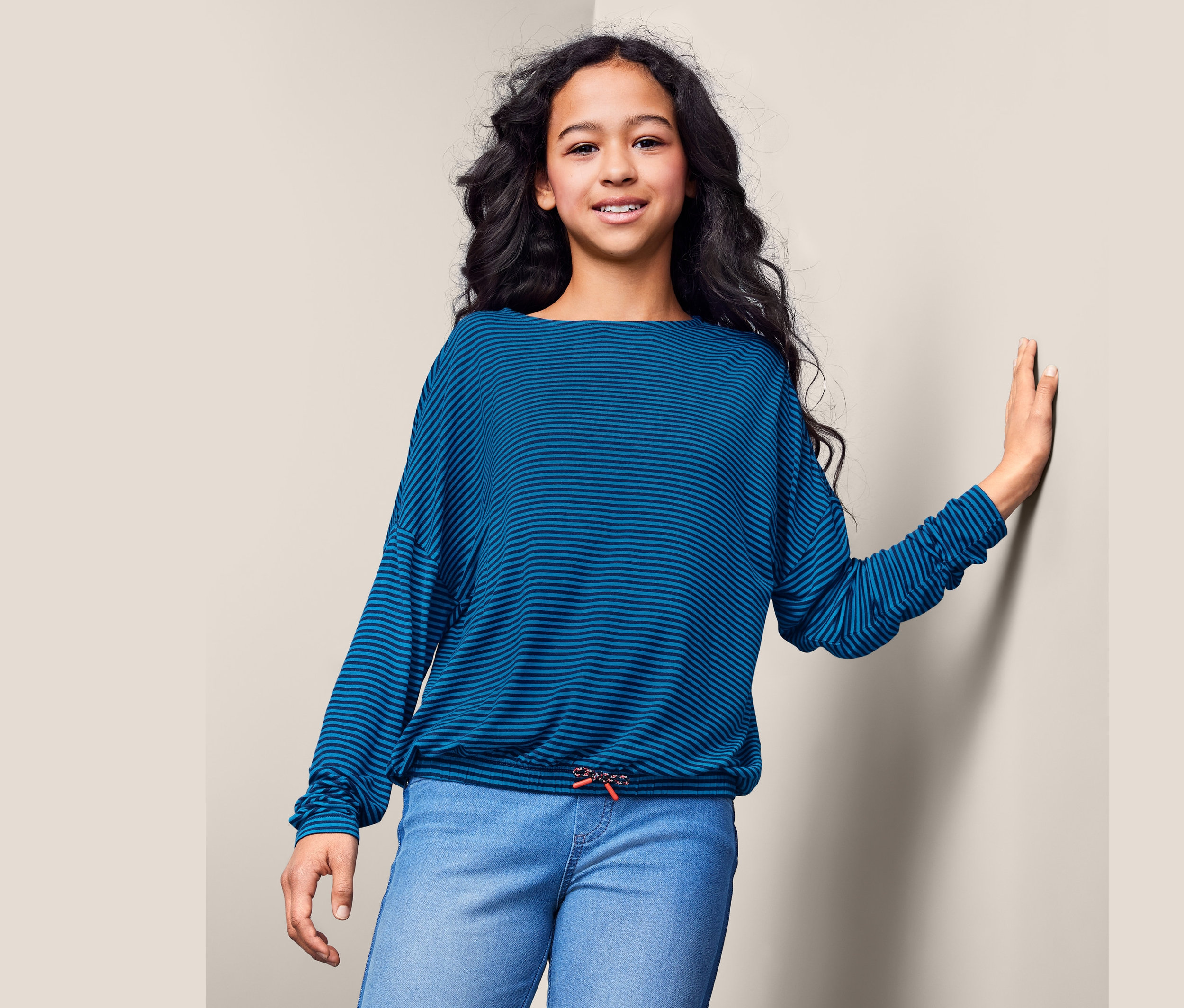 Une fille appuie sa main contre le mur et porte un haut à manches longues rayé bleu et un jean.