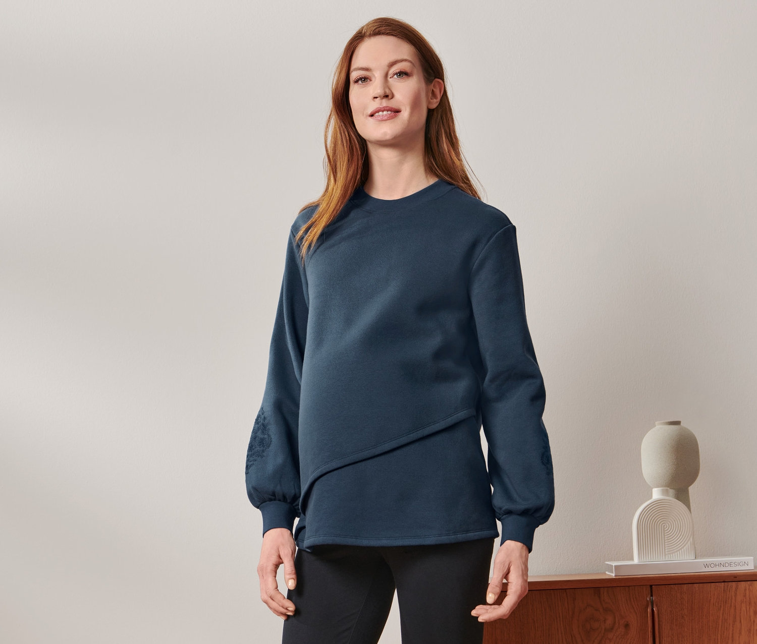 Une femme enceinte debout portant un sweat-shirt de grossesse et d'allaitement, un pantalon noir et des baskets.