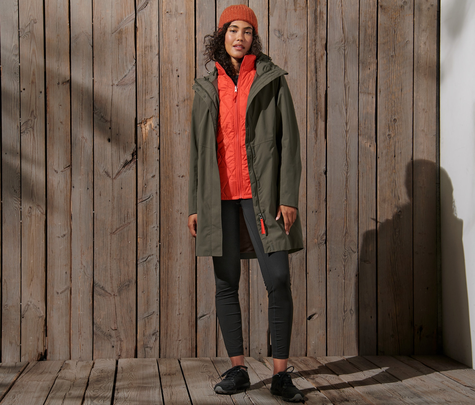 Une femme porte un manteau de pluie 3-en-1 olive avec un gilet orange et un bonnet orange.