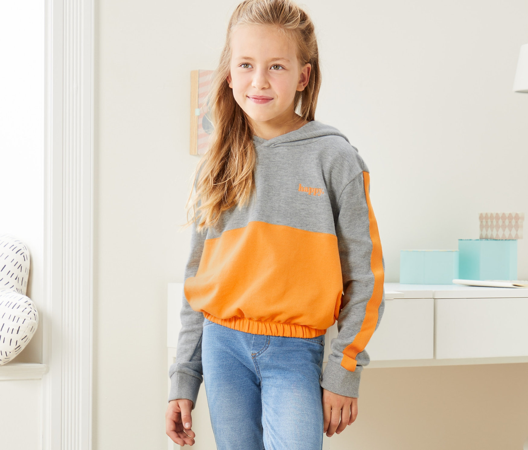 Une fille porte un sweat-shirt court à capuche gris-jaune chiné, un jean et des baskets.