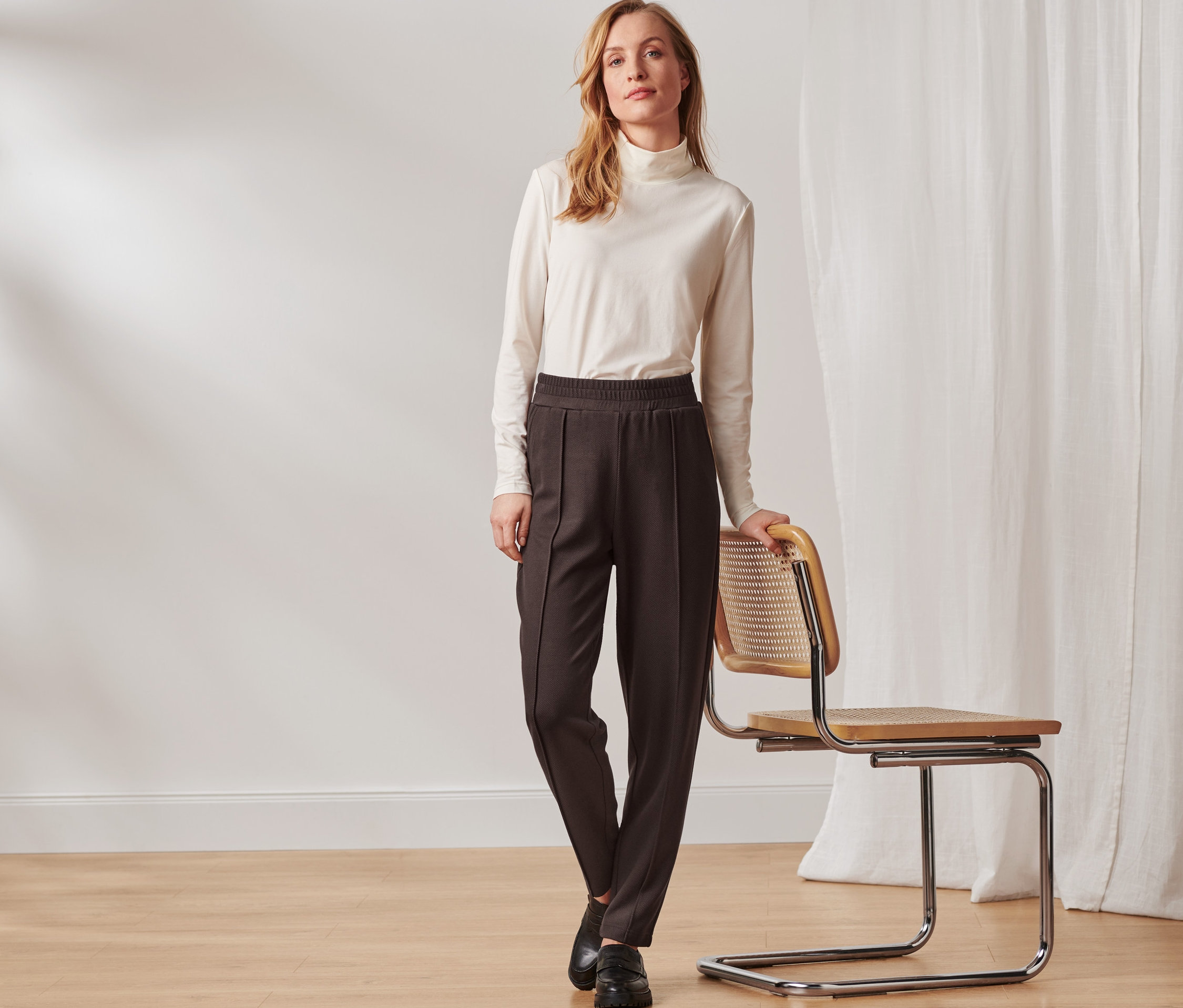 Une femme pose à côté d'une chaise et porte un pantalon de jogging marron foncé avec une nervure.