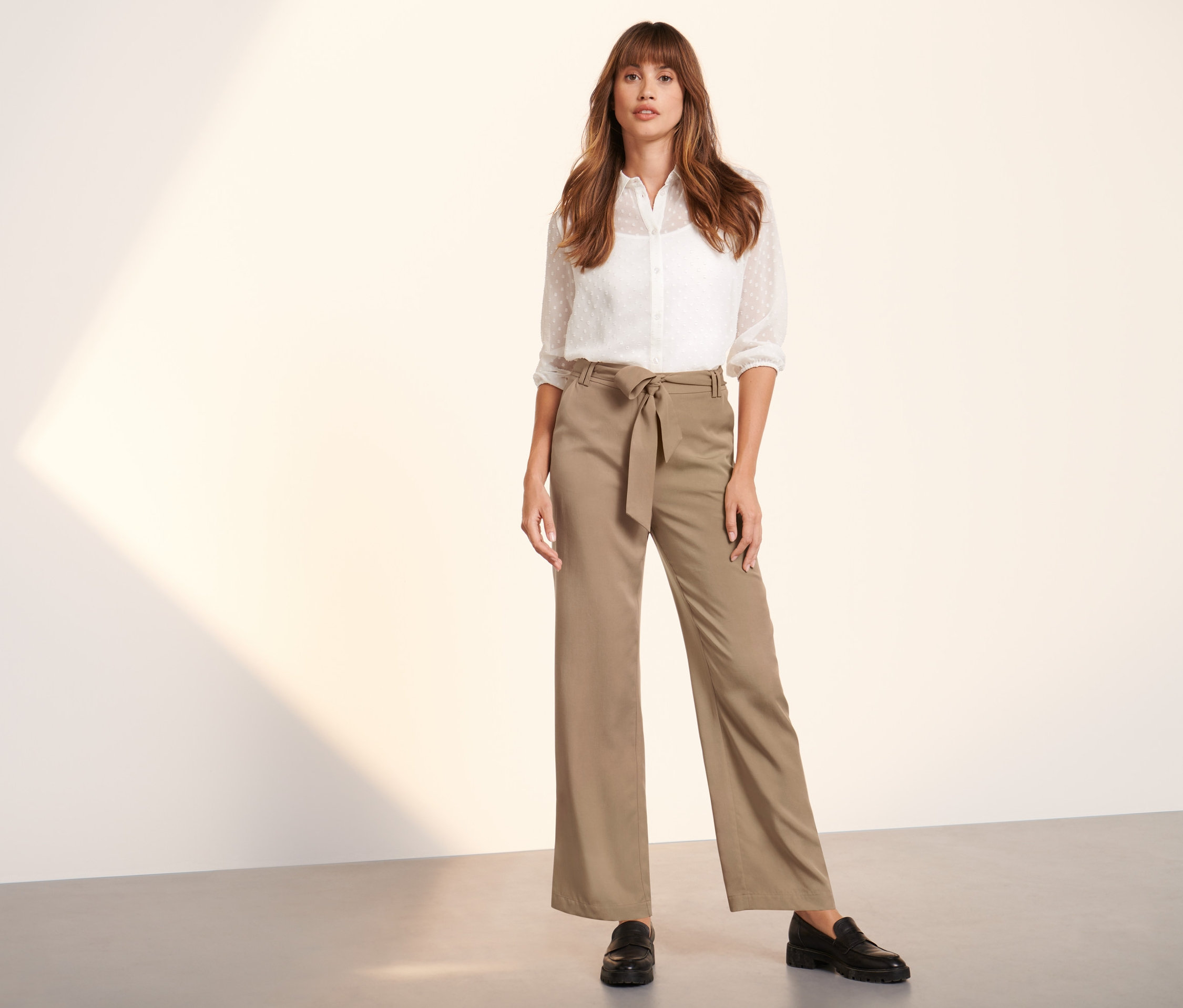 Une femme pose, portant un pantalon tissé beige avec ceinture et une chemise blanche.