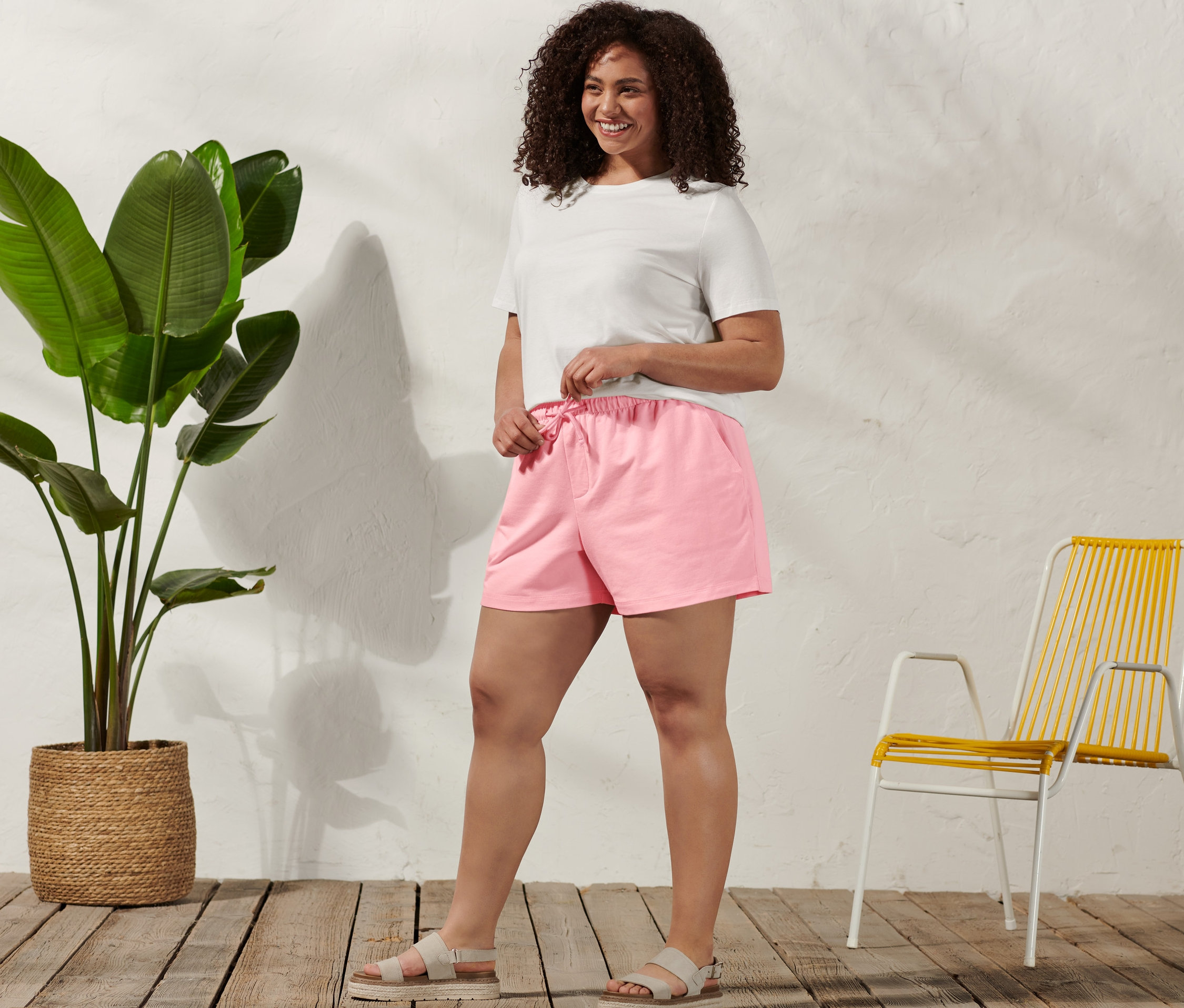 Une femme debout serre le cordon de son short molletonné, elle porte un t-shirt basique, blanc, et des sandales confortables.