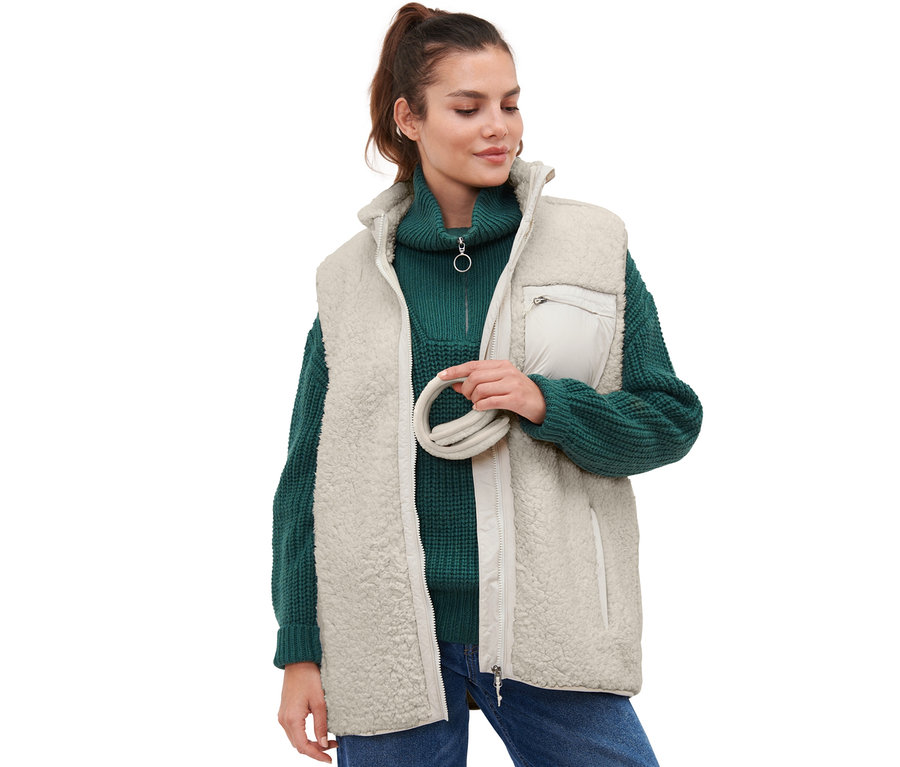 Une femme tient un cache-oreilles couleur crème et porte un pull vert et un gilet crème.