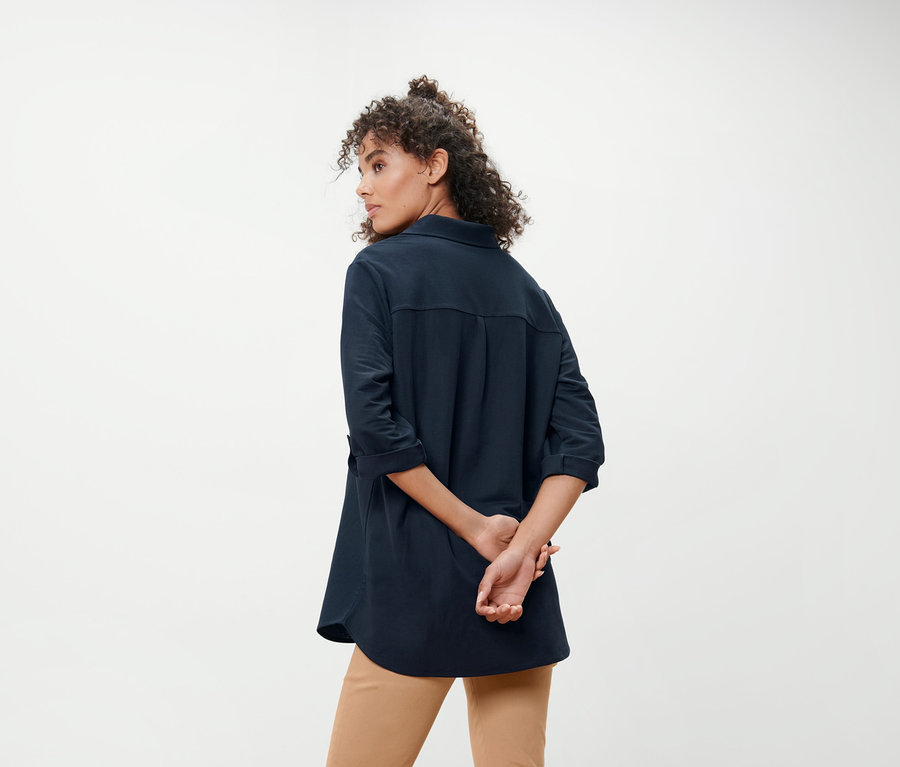 Une femme se tient dos à la caméra, les mains derrière le dos. Elle porte un chemisier en jersey piqué bleu foncé et un pantalon kaki.