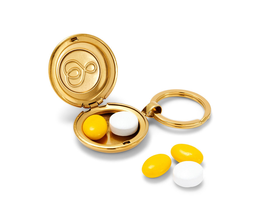 Un porte-clés boîte à pilules doré ouvert contient une pilule jaune et une blanche, deux pilules sont posées à côté.