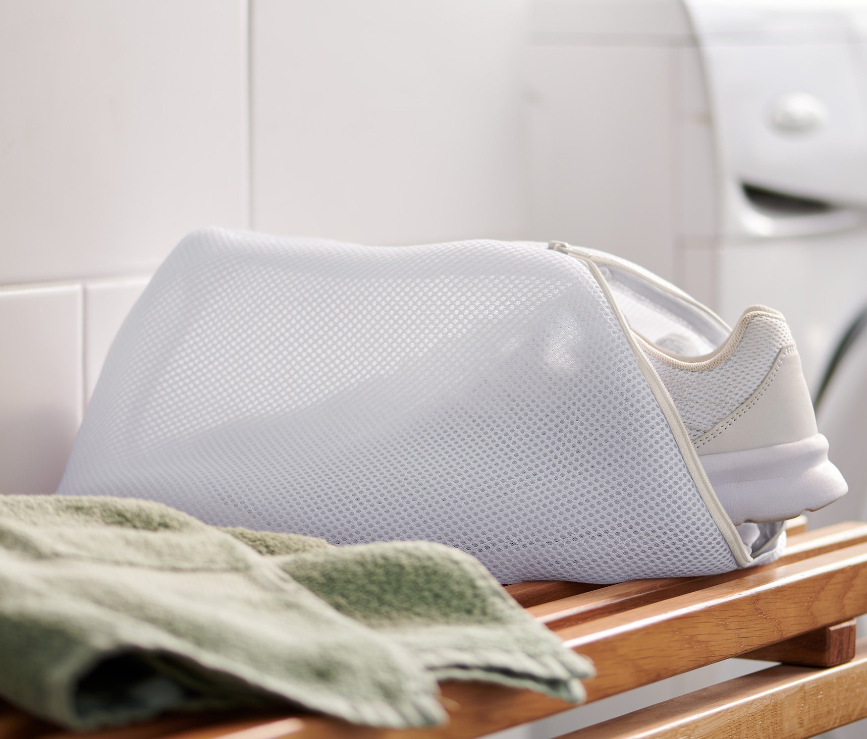 Une basket blanche est placée dans un filet de lavage pour chaussures, posé sur un banc en bois à côté d'une serviette verte. Une machine à laver blanche et des carreaux blancs sont visibles en arrière-plan.