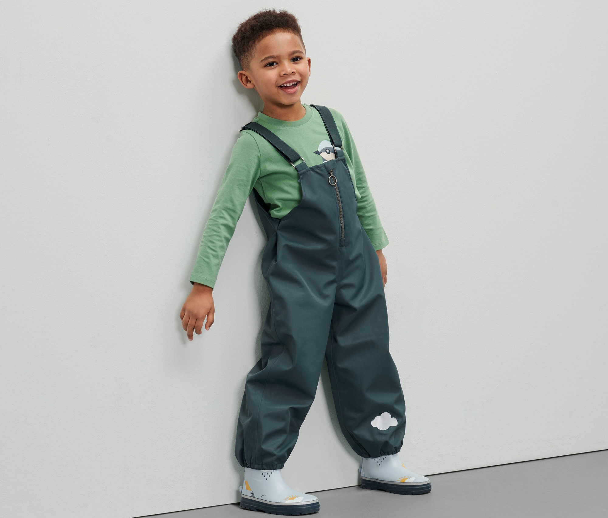 Un garçon portant un pantalon imperméable pour tout petit est appuyé contre un mur.