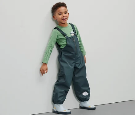 Un garçon portant un pantalon imperméable pour tout petit est appuyé contre un mur.