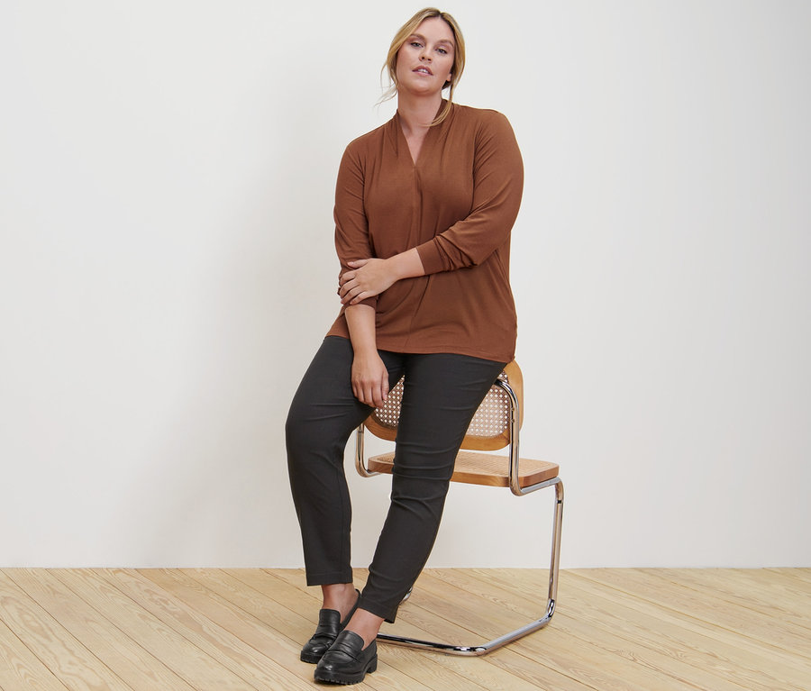 Une femme est assise sur une chaise et porte un t-shirt manches longues avec encolure en V couleur cognac, un pantalon en bengaline et des loafers en cuir.