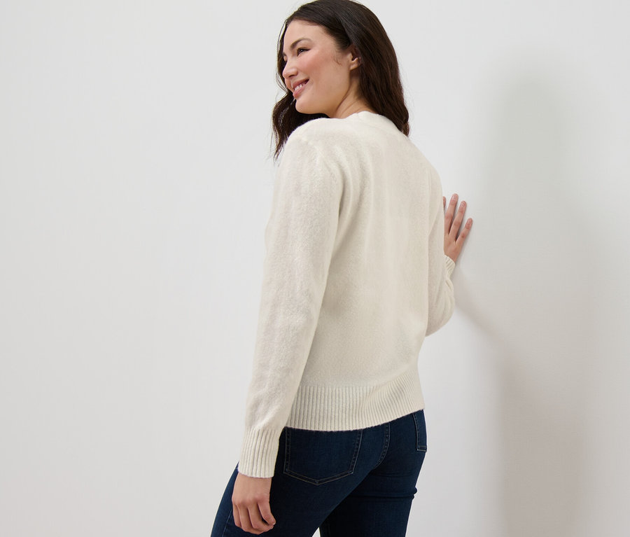 Une femme regarde par-dessus son épaule et s'appuie contre un mur en portant un cardigan blanc crème à structure tricotée.