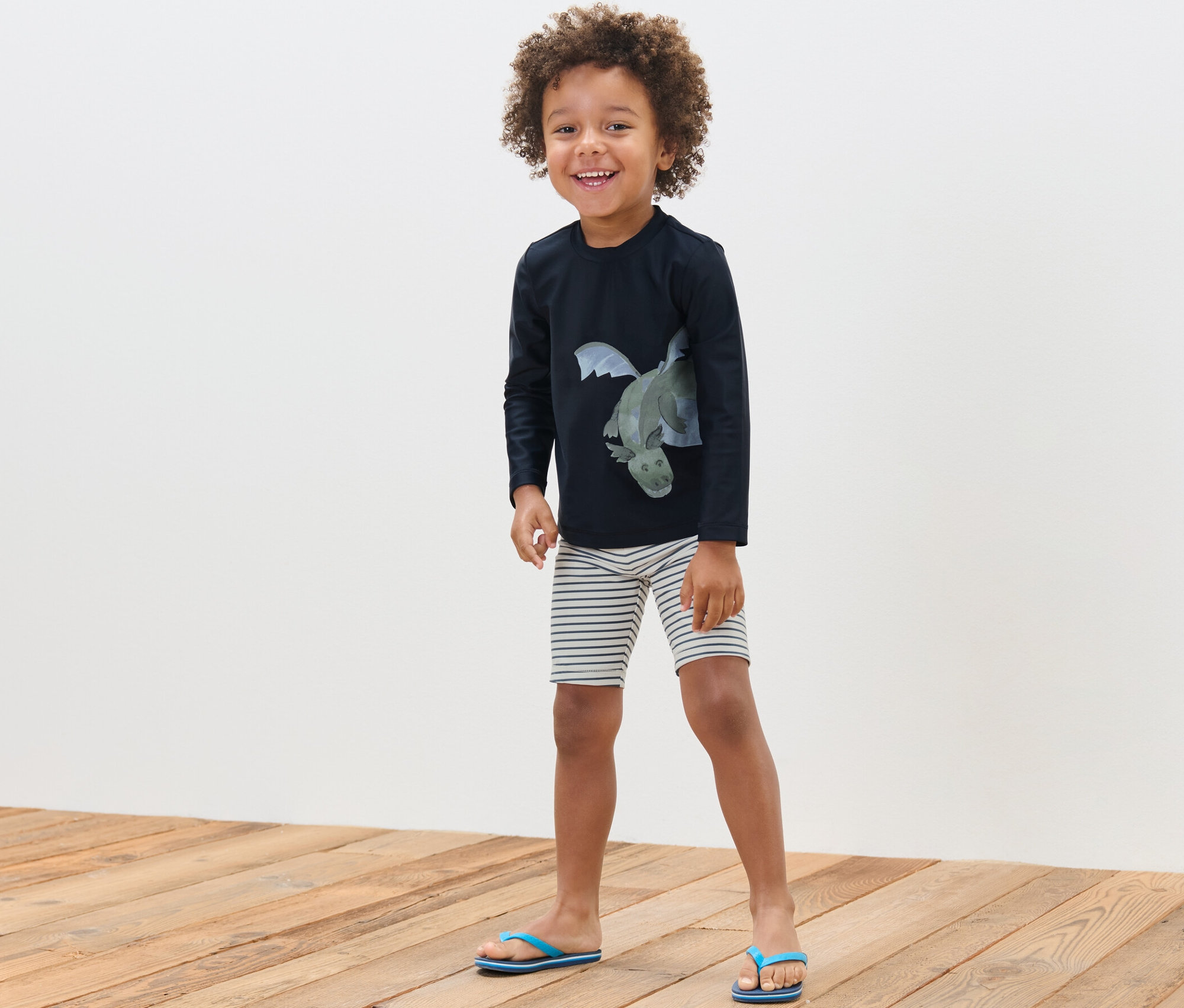 Un garçon souriant pose, portant un ensemble pour enfant avec indice de protection UV 80, dragon.