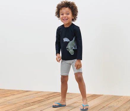 Un garçon souriant pose, portant un ensemble pour enfant avec indice de protection UV 80, dragon.