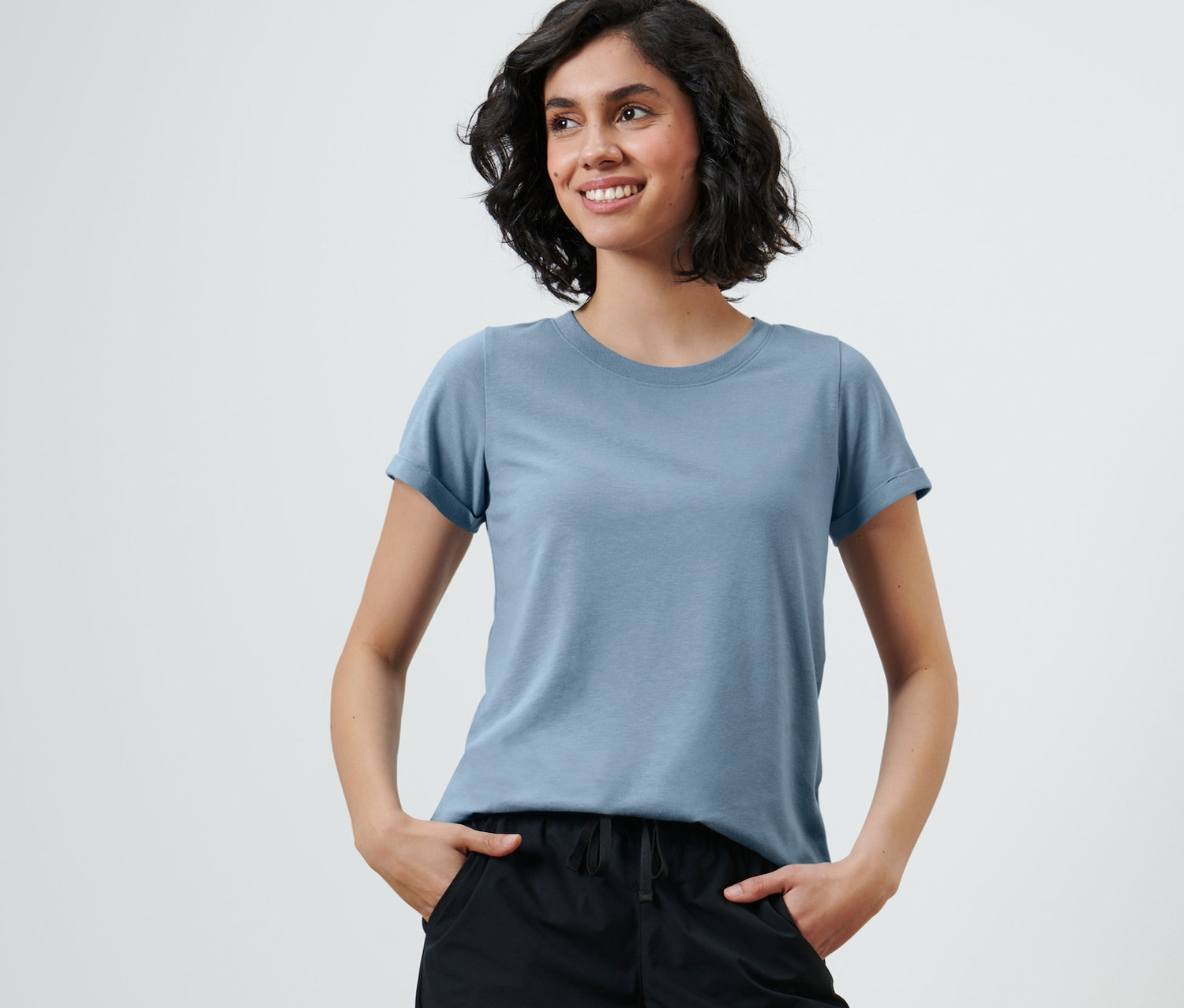 Femme souriante avec les mains dans les poches d'un pantalon noir et un t-shirt technique.