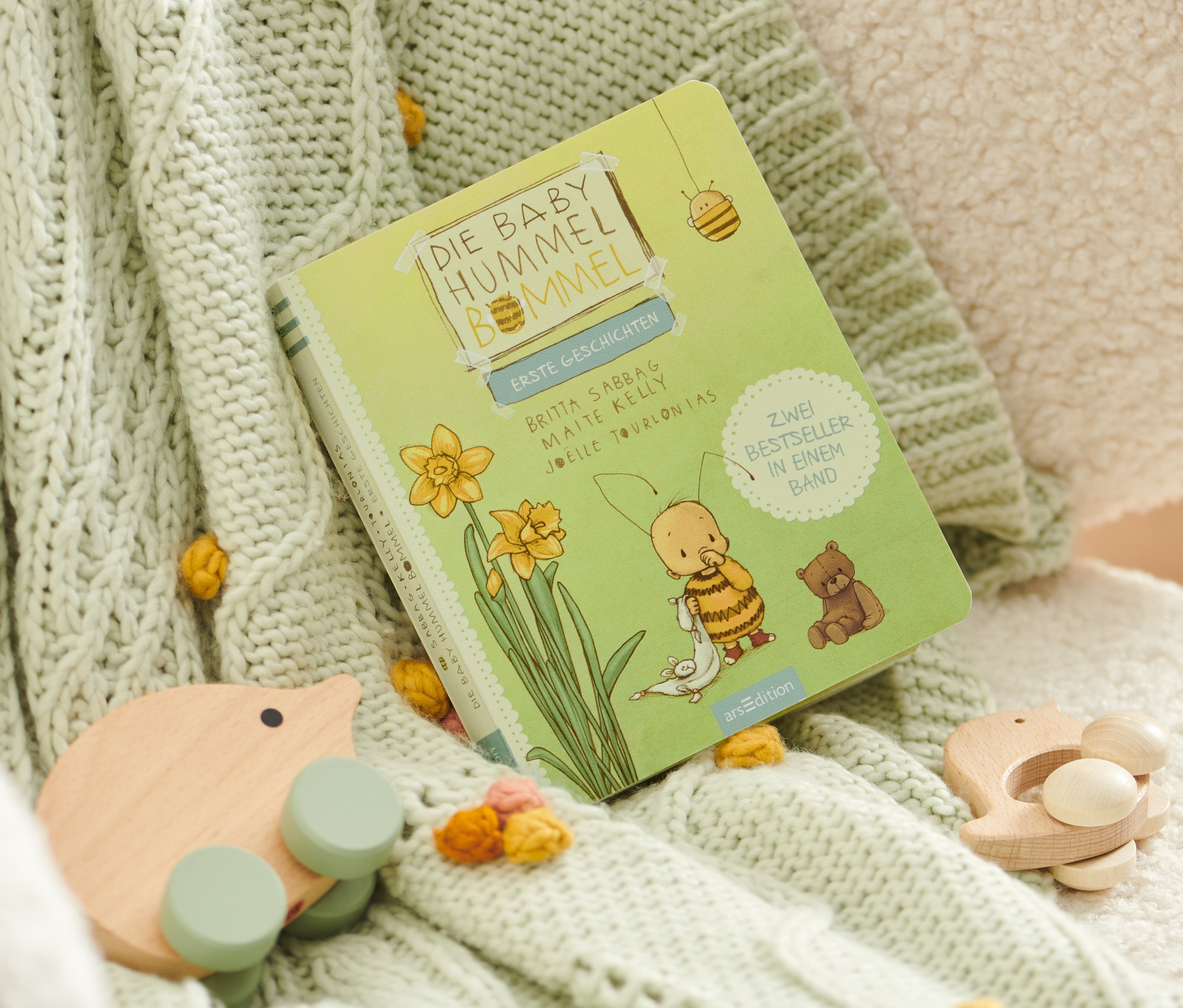 Le livre « Baby Hummel Bommel – erste Geschichten » est posé sur une couverture tricotée avec des pompons et des jouets en bois.